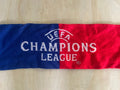 A knitted Champions League scarf with a vertical split design in blue and red, featuring white embroidered text and the UEFA logo, laid flat on a light wood surface.