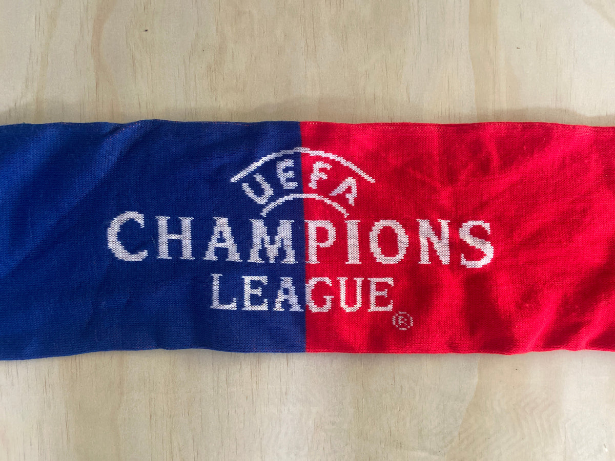 A knitted Champions League scarf with a vertical split design in blue and red, featuring white embroidered text and the UEFA logo, laid flat on a light wood surface.