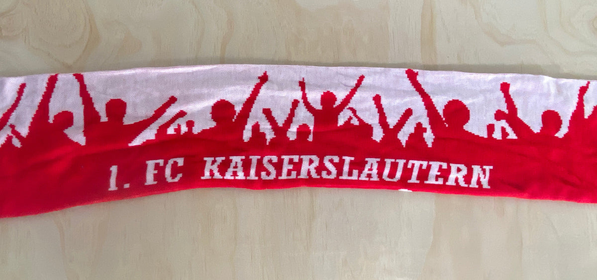 A red and white scarf featuring the text '1. FC KAISERSLAUTERN' in white on a red background, with red silhouettes of cheering fans along the top edge, laid flat on a light wood surface.