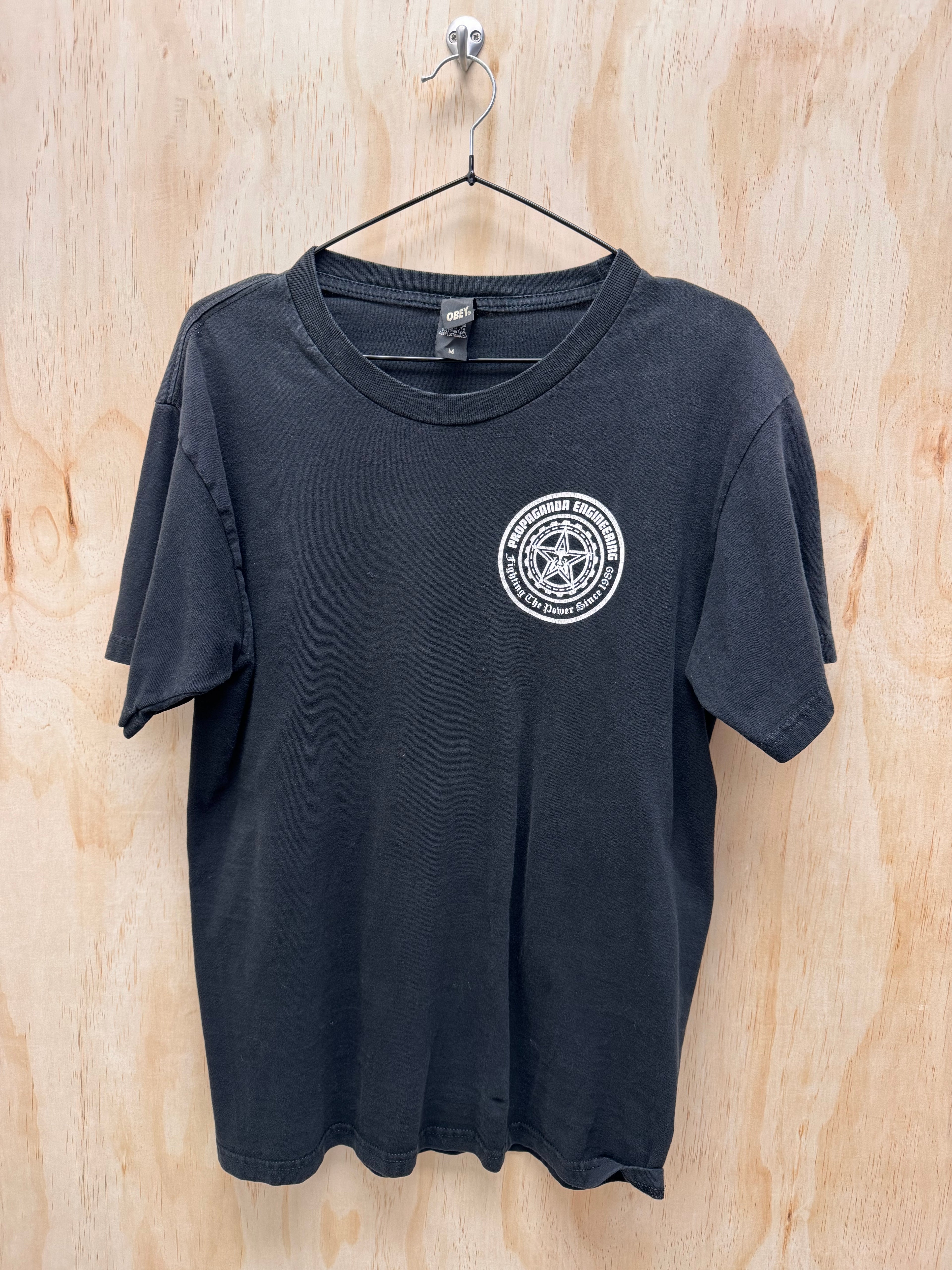 A black short-sleeve t-shirt hanging on a black wire hanger against a light wood panel background. The shirt features a white circular graphic printed on the left chest containing a star design and the text 'SOUTH CAROLINA DEFENSE FORCE'.