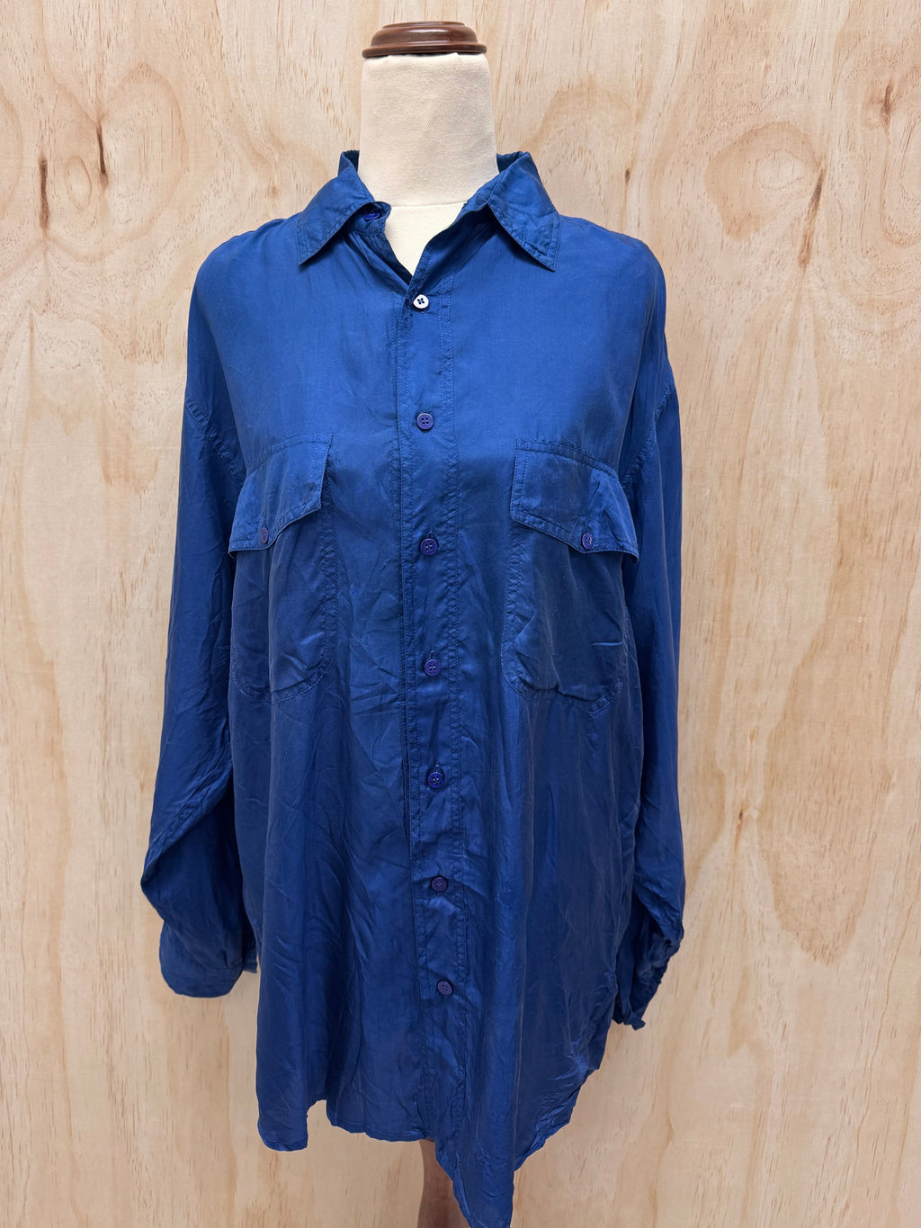 A blue long-sleeve button-up shirt with two chest flap pockets, displayed on a mannequin torso against a light wood plywood background.