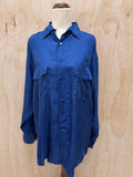 A blue long-sleeve button-up shirt with two chest flap pockets, displayed on a mannequin torso against a light wood plywood background.