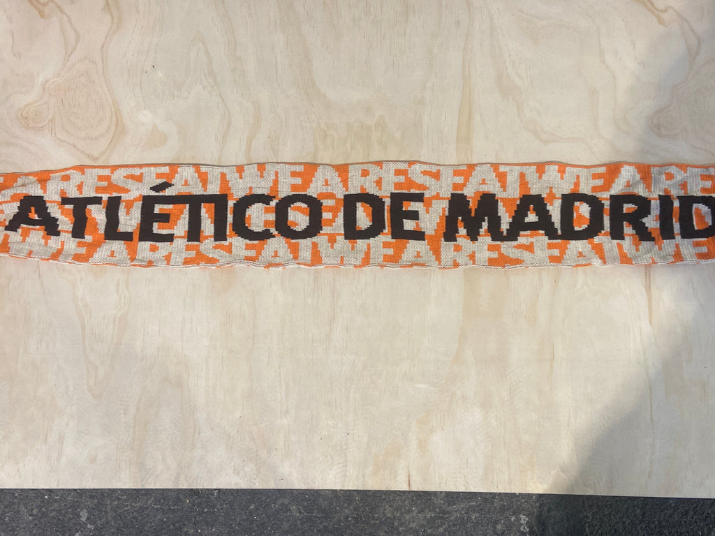 A close-up of a white Atlético de Madrid scarf featuring the club's orange and black branding, laid flat on a light wood background.