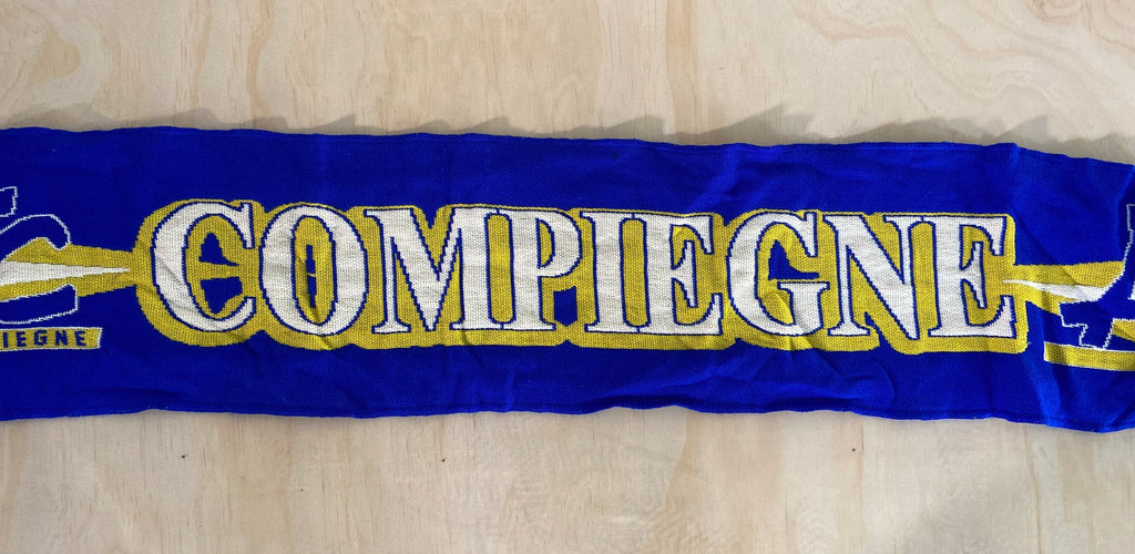 A royal blue knit scarf with 'COMPIEGNE' written in white and gold, laid flat on a light wood background.