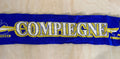 A royal blue knit scarf with 'COMPIEGNE' written in white and gold, laid flat on a light wood background.