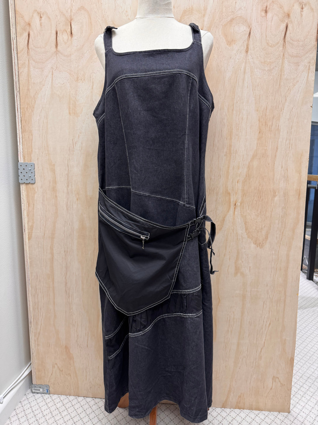 A dark washed denim dress displayed on a mannequin torso in front of a plywood backdrop. The sleeveless dress features contrast light stitching, a square neckline, a draped asymmetrical front panel, and a side waist tie.
