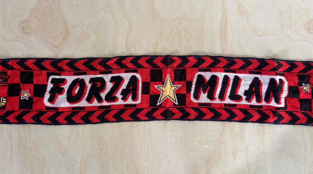 A red and black striped knit scarf with 'FORZA MILAN' embroidered in white and black, featuring a gold star in the center, laid flat on a light wood background.