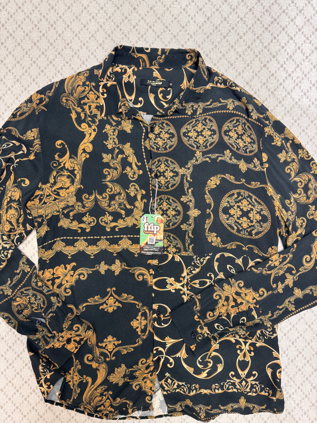 A long-sleeve button-up shirt with a black background and an ornate gold baroque-style damask pattern, laid flat on a textured light background. A green and white 'hip' brand tag is attached to the collar.