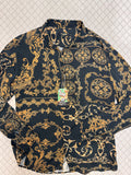 A long-sleeve button-up shirt with a black background and an ornate gold baroque-style damask pattern, laid flat on a textured light background. A green and white 'hip' brand tag is attached to the collar.