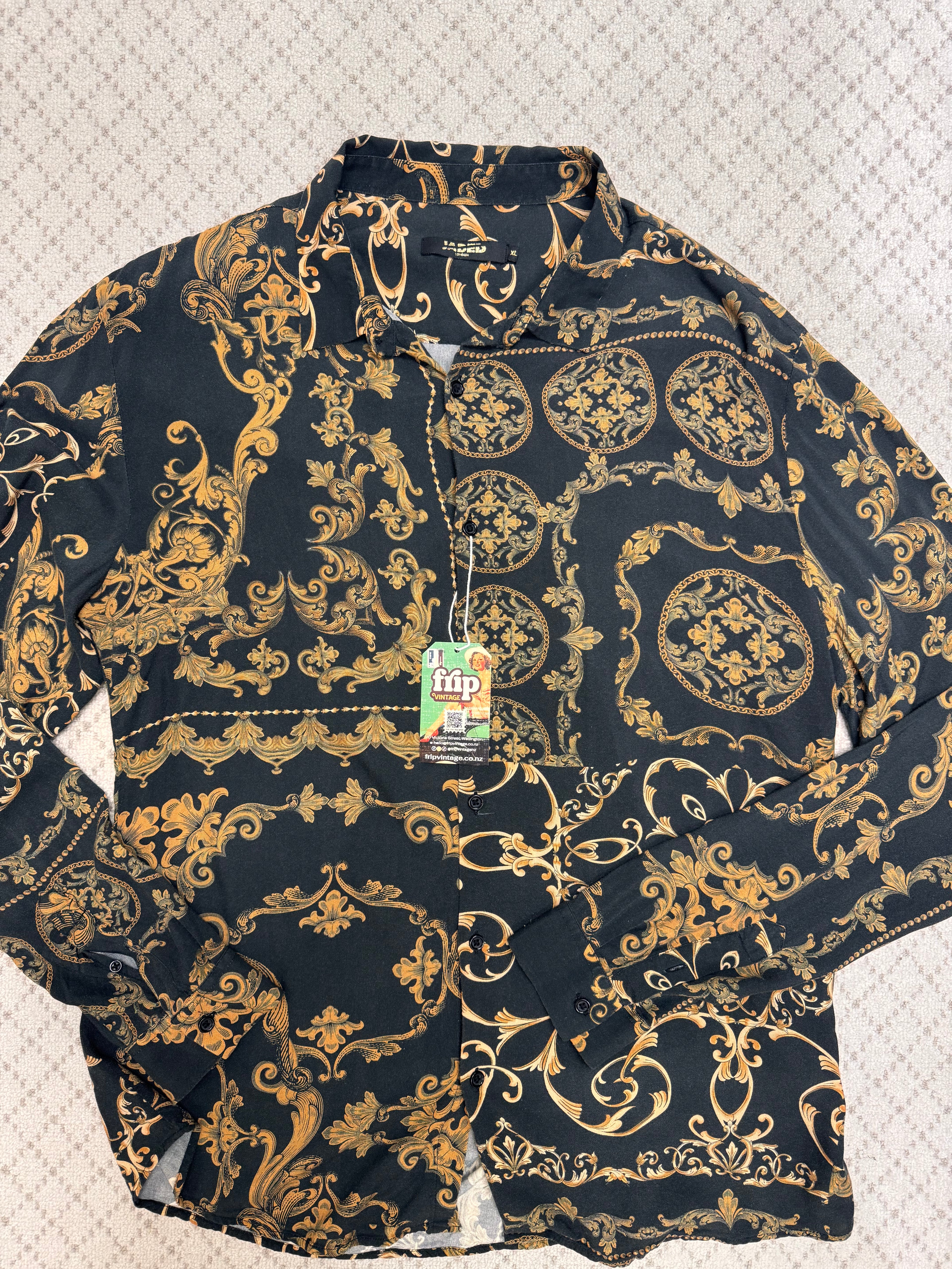 A long-sleeve button-up shirt with a black background and an ornate gold baroque-style damask pattern, laid flat on a textured light background. A green and white 'hip' brand tag is attached to the collar.