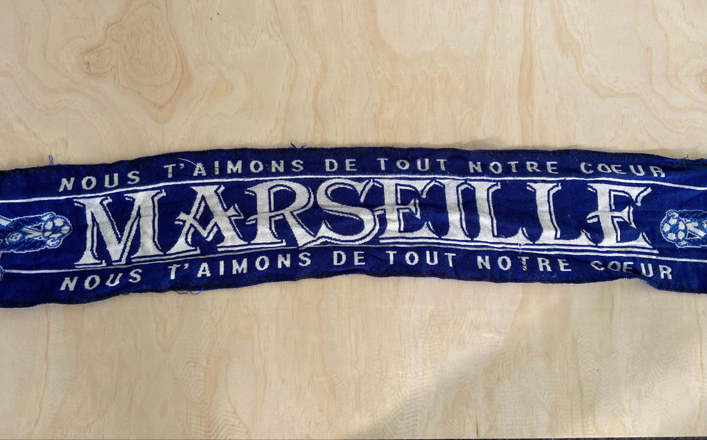 A dark blue fabric banner or scarf with white printed text reading 'MARSILIE' and surrounding French text, laid flat on a light wood surface.