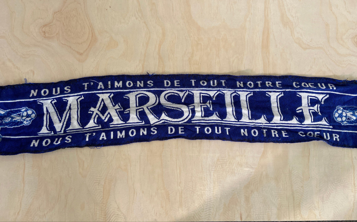 A dark blue fabric banner or scarf with white printed text reading 'MARSILIE' and surrounding French text, laid flat on a light wood surface.