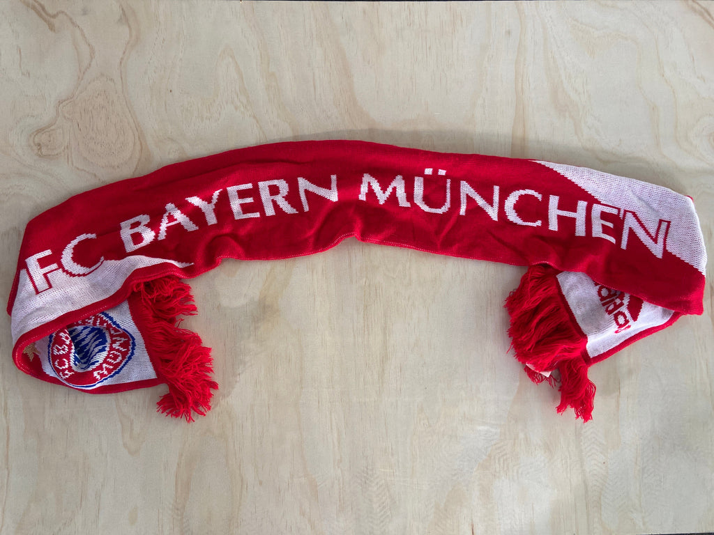 A red and white knit scarf with 'FC BAYERN MÜNCHEN' written across it, laid flat on a light wood surface.