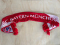 A red and white knit scarf with 'FC BAYERN MÜNCHEN' written across it, laid flat on a light wood surface.