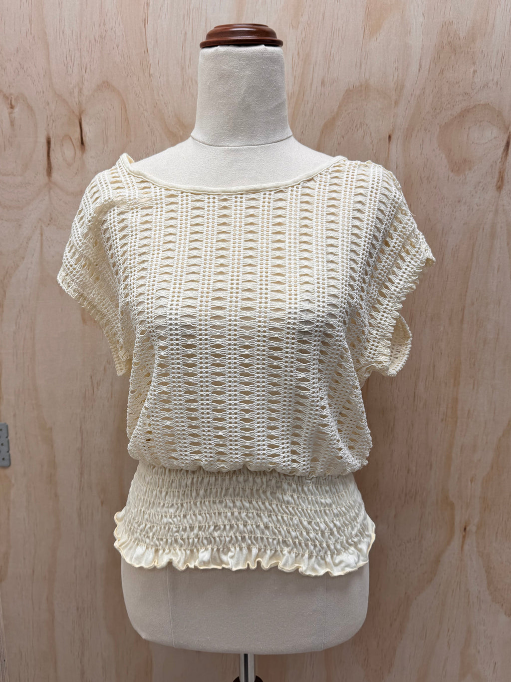 An off-the-shoulder cream-colored knit top with a smocked ruffled hem, displayed on a dress form mannequin against a light wood panel background.