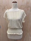 An off-the-shoulder cream-colored knit top with a smocked ruffled hem, displayed on a dress form mannequin against a light wood panel background.