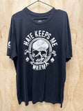 A black graphic t-shirt hanging on a black hanger against a light wood grain background. The shirt features a large white graphic on the front with a skull, crossed tools, and the text 'HATE KEEPS ME WARM' in an arc.