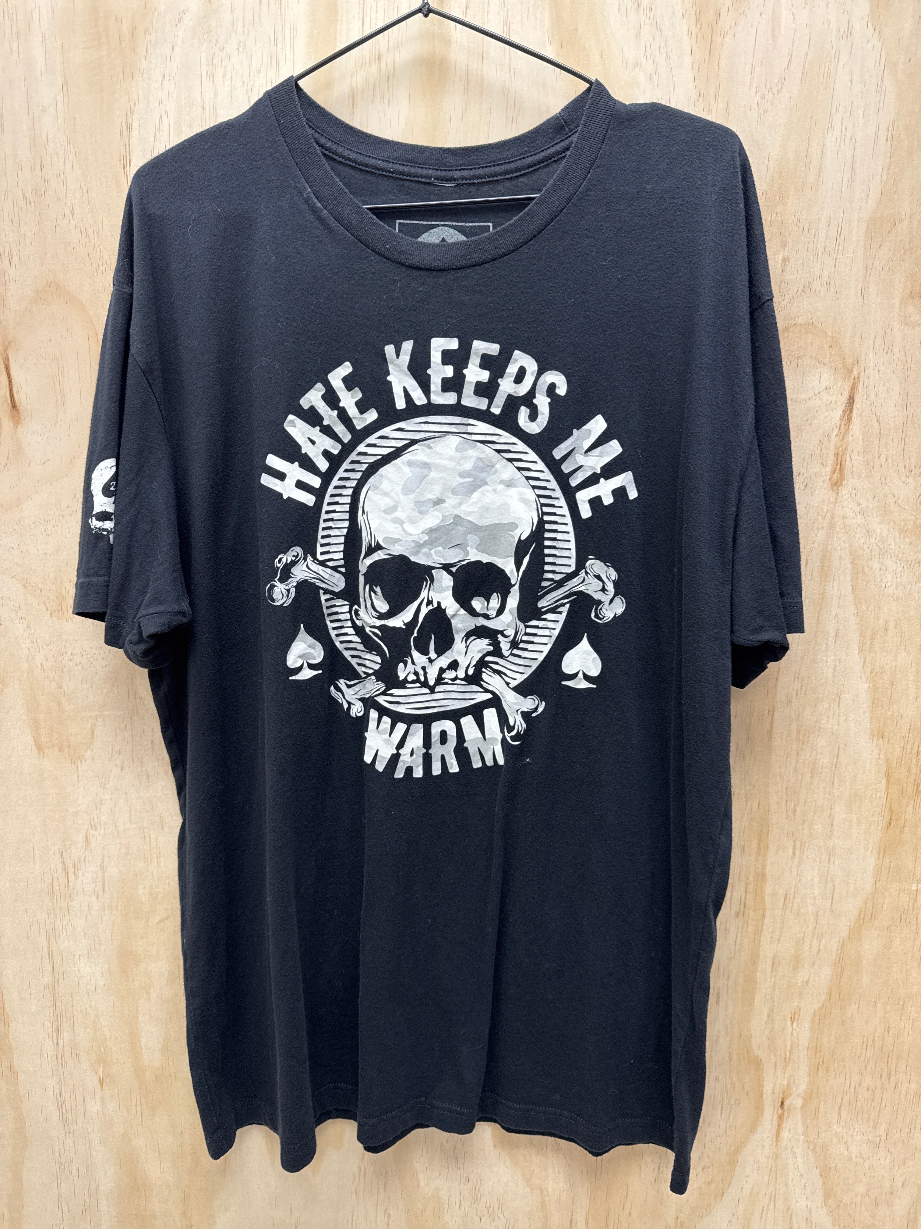 A black graphic t-shirt hanging on a black hanger against a light wood grain background. The shirt features a large white graphic on the front with a skull, crossed tools, and the text 'HATE KEEPS ME WARM' in an arc.