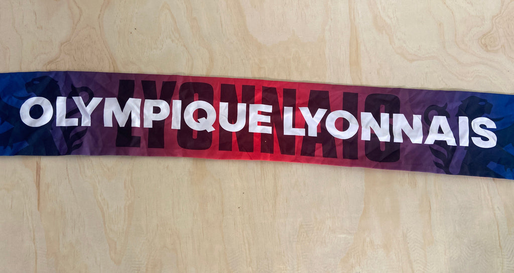 A horizontal athletic scarf with 'OLYMPIQUE LYONNAIS' printed in bold white letters over a red-to-purple gradient and dark side patterns, laid flat on a light wood board.