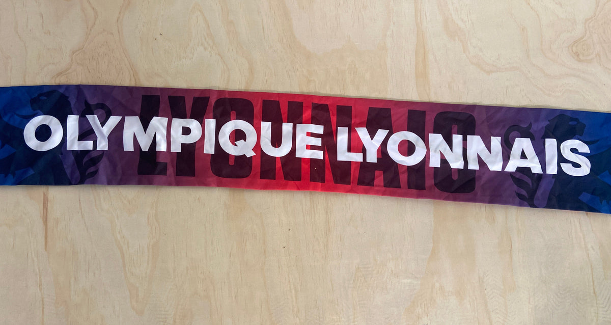 A horizontal athletic scarf with 'OLYMPIQUE LYONNAIS' printed in bold white letters over a red-to-purple gradient and dark side patterns, laid flat on a light wood board.