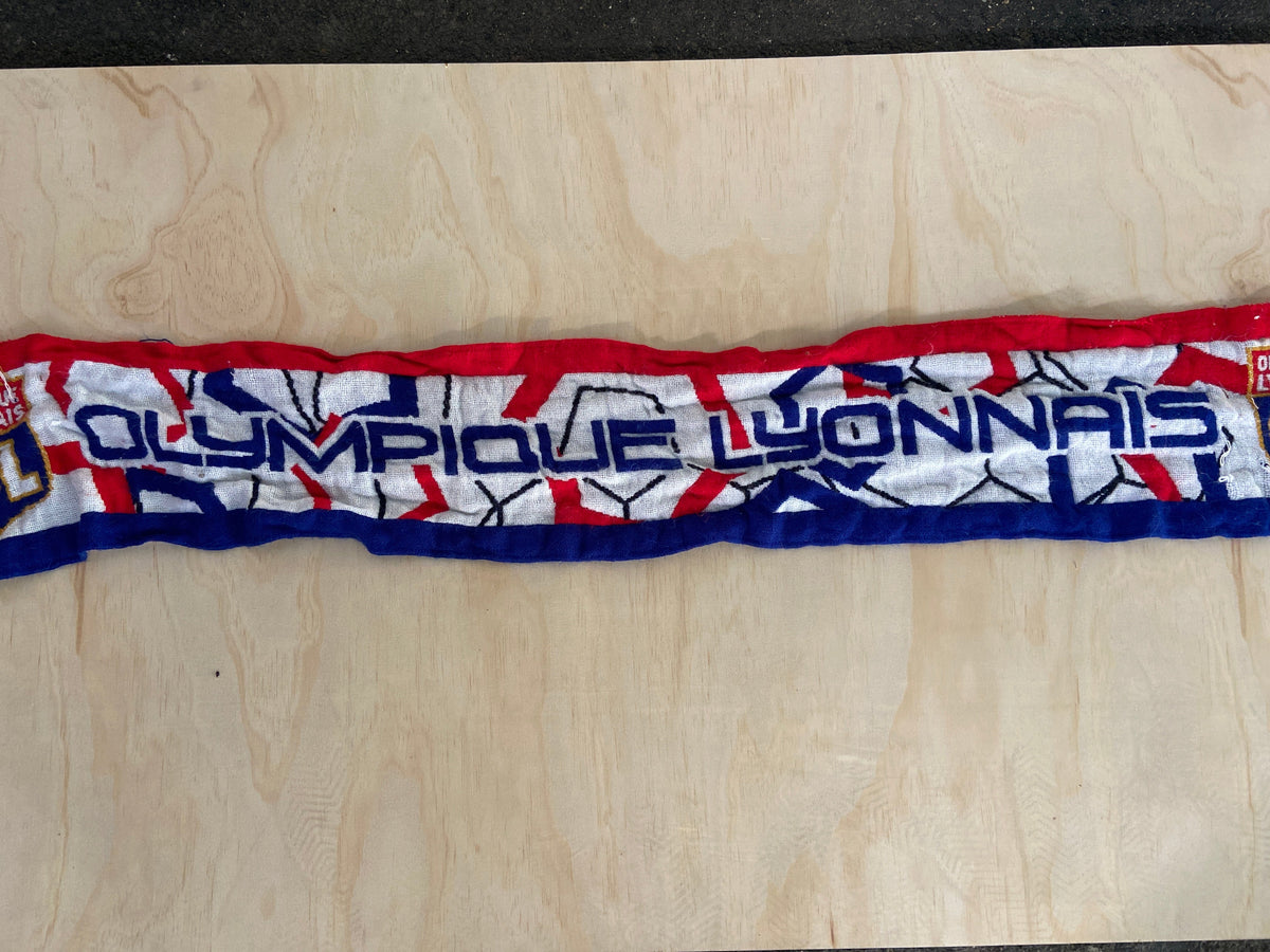 A vertical close-up of a long, narrow scarf laid flat on a light wood surface. The scarf has a white base with bold blue and red graphics and large vertical text reading 'Olympique Lyonnais'
