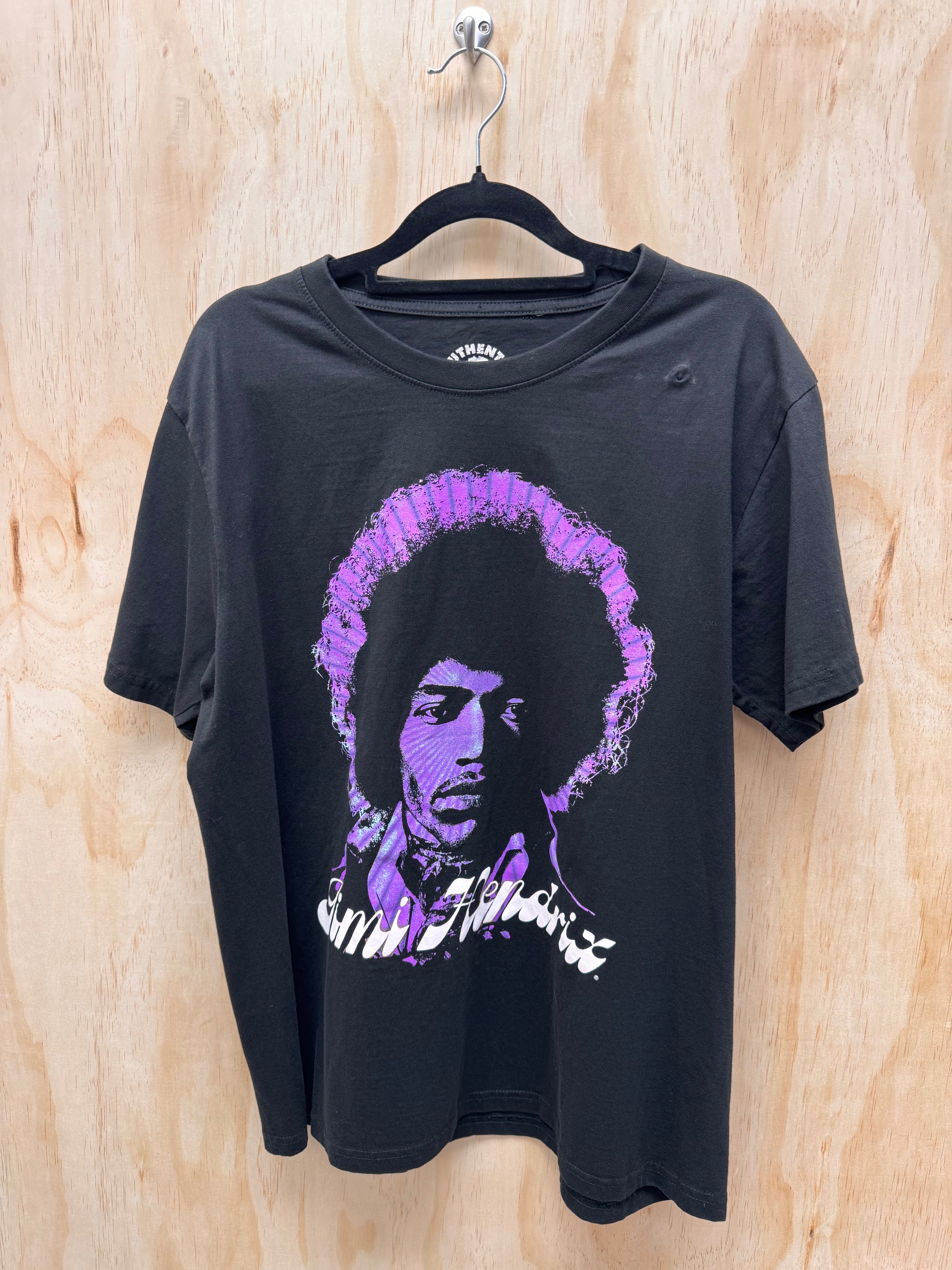 A black short-sleeve graphic t-shirt hanging on a black hanger against a light wood panel background. The shirt features a stylized purple and white graphic portrait of Jim Morrison on the front.